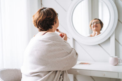 Smiling middle aged plus size woman doing makeup near mirror at home