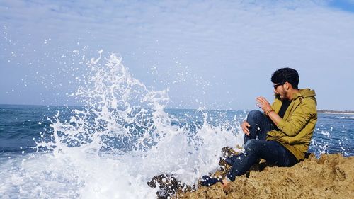 Side view of young man sitting on rock by sea against cloudy sky