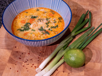 Asian coconut soup with red curry paste in a blue bowl on a wooden block with spring onions, lime.