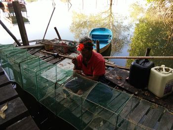 High angle view of man sewing fishing net on wooden plank over lake
