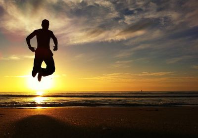 Full length rear view of silhouette man jumping into sea on beach at sunset