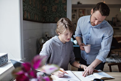 Father holding coffee cup while assisting son in studying at home