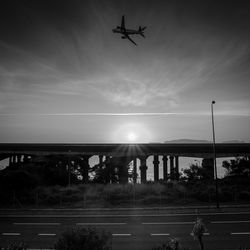 Silhouette airplane flying over street against sky during sunset