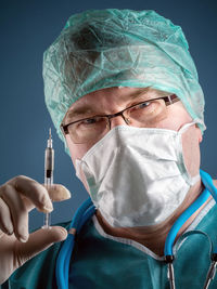 Close-up portrait of doctor wearing surgical cap and mask while holding syringe against wall