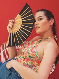 Young woman with hand fan looking away while sitting on chair against red wall