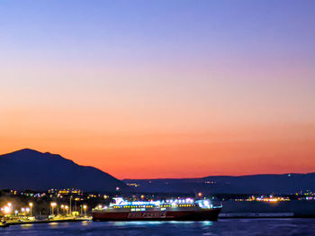 Illuminated city by sea against sky at sunset
