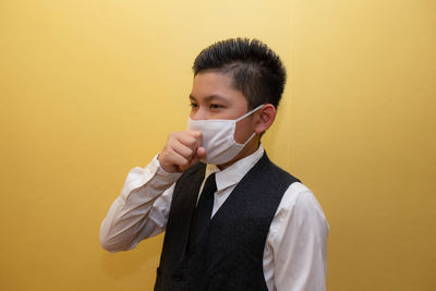 Boy wearing mask coughing while standing against yellow wall