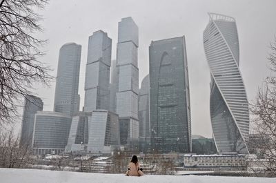 Rear view of woman looking at modern buildings while sitting on snow covered park