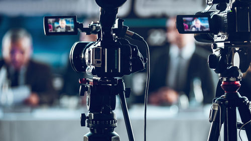 Two cameras recording presentation at press conference, blurred speakers wearing suit background