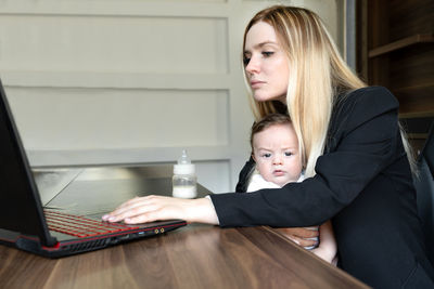 Young businesswoman holds baby in her arms, in office, combining this with working on laptop.