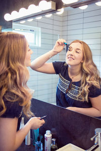 Happy young woman applying mascara in front of bathroom mirror