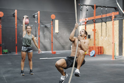 Young man exercising with rope while girlfriend lifting kettlebell in gym