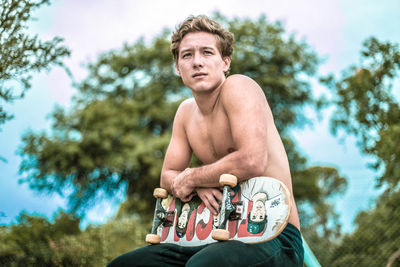 Portrait of shirtless young man holding plant against trees