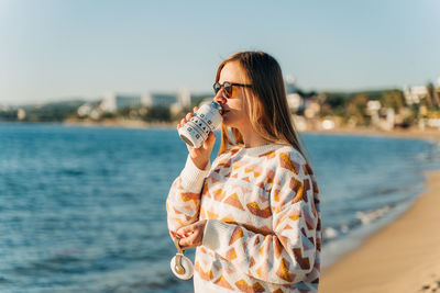 Young woman drinking water while standing against sea