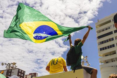 Low angle view of man holding brazilian flag by friend against buildings on sunny day