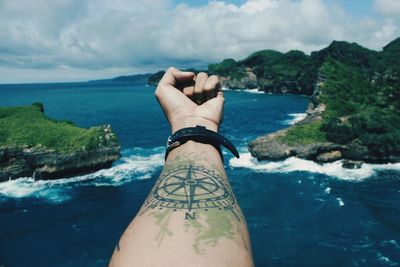 Cropped hand with tattoo by sea against cloudy sky