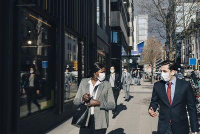 Male and female colleagues discussing while walking on sunny day in city during covid-19