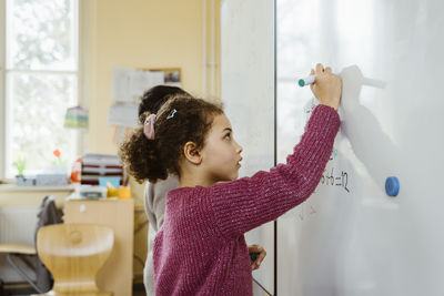 Side view of girl and boy writing on whiteboard in classroom