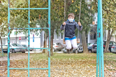 Small kid with face mask swinging at the playground during coronavirus pandemic.