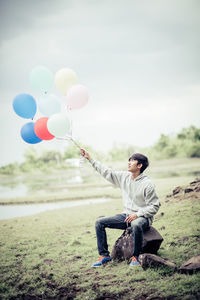 Full length of man holding balloons while sitting on rock against sky