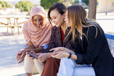 Female friends sharing smart phone sitting on bench