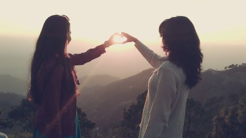 Female friends making heart shape from hands in front of sun against sky