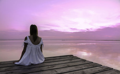 Rear view of woman sitting on shore against sunset sky