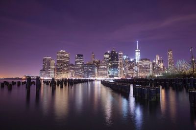 Wooden posts in east river by illuminated city against sky