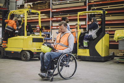 Full length of male carpenter with disability using digital tablet sitting on wheelchair in warehouse