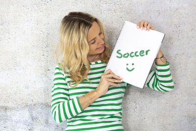 Smiling beautiful woman holding book with text while standing against wall