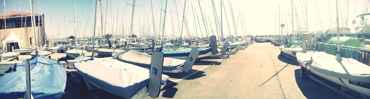 nautical vessel, transportation, moored, boat, mode of transport, in a row, water, sunlight, harbor, sea, built structure, day, panoramic, architecture, mast, sky, outdoors, no people, clear sky, rope