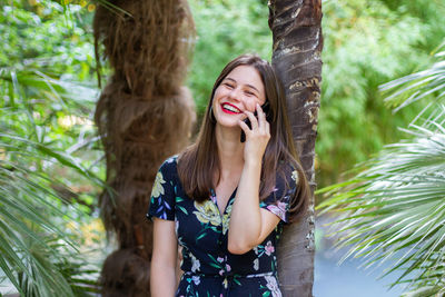 Smiling young woman using phone while standing against trees