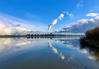 An industrial area with a plume of smoke and clouds above is reflected in a perfectly smooth lake