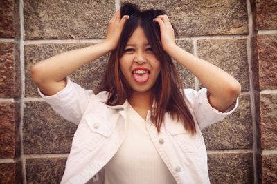 Portrait of young woman with hands in hair sticking out tongue against wall