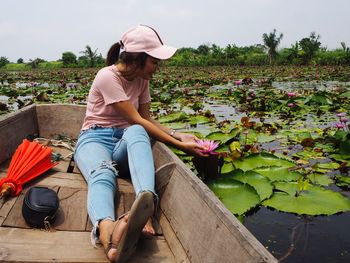 Full length of young woman touching lotus water lily while sitting in boat against sky