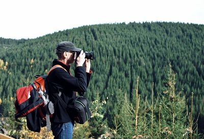 Side view of man photographing against trees and sky