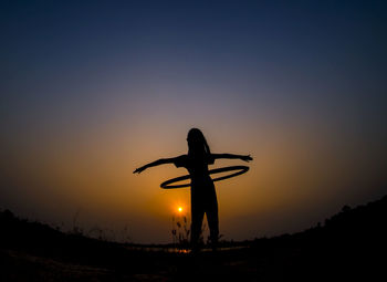 Silhouette girl playing with hula hoop crane against orange sky