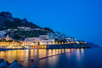 Illuminated buildings by sea against clear blue sky at night