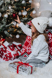 Happy little girl in white hat holding christmas gift boxes while sitting on floor.