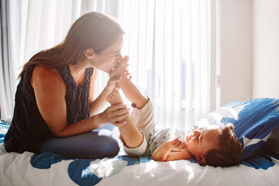 Young caucasian mother kissing boy toddler son feet heels legs. mom playing with child baby on bed 