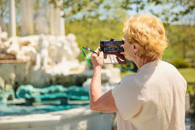 Elderly woman photographing on a mobile phone a fountain in the park