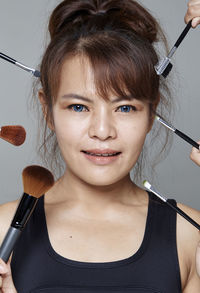 Close-up portrait of smiling beautiful woman with various make-up brushes and mascara applicator against gray background