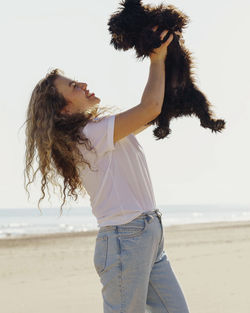 Side view of woman on the beach raising her dog to the sky