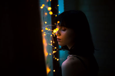 Close-up of woman with illuminated string lights on window at home