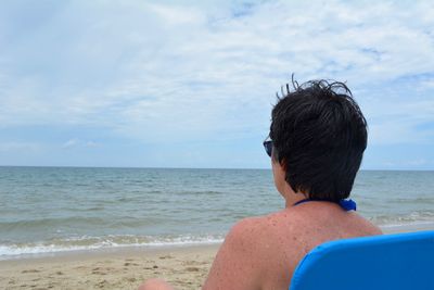 Rear view of mature woman looking at sea while sitting on deck chair against sky