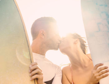 Low angle view of couple with surfboards kissing against sky during sunny day