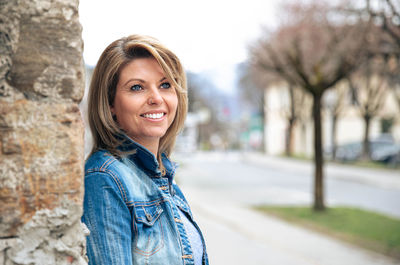 Radiant woman in denim smiles warmly, leaning against textured wall