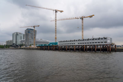 Cranes by sea against sky in city