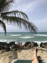 Sandy feet on beach against sky with palm leaves