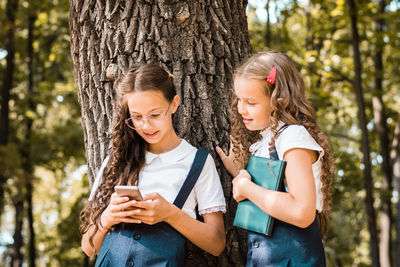 Smiling pupils in school uniform with a book looking at a smartphone in the park on a warm day